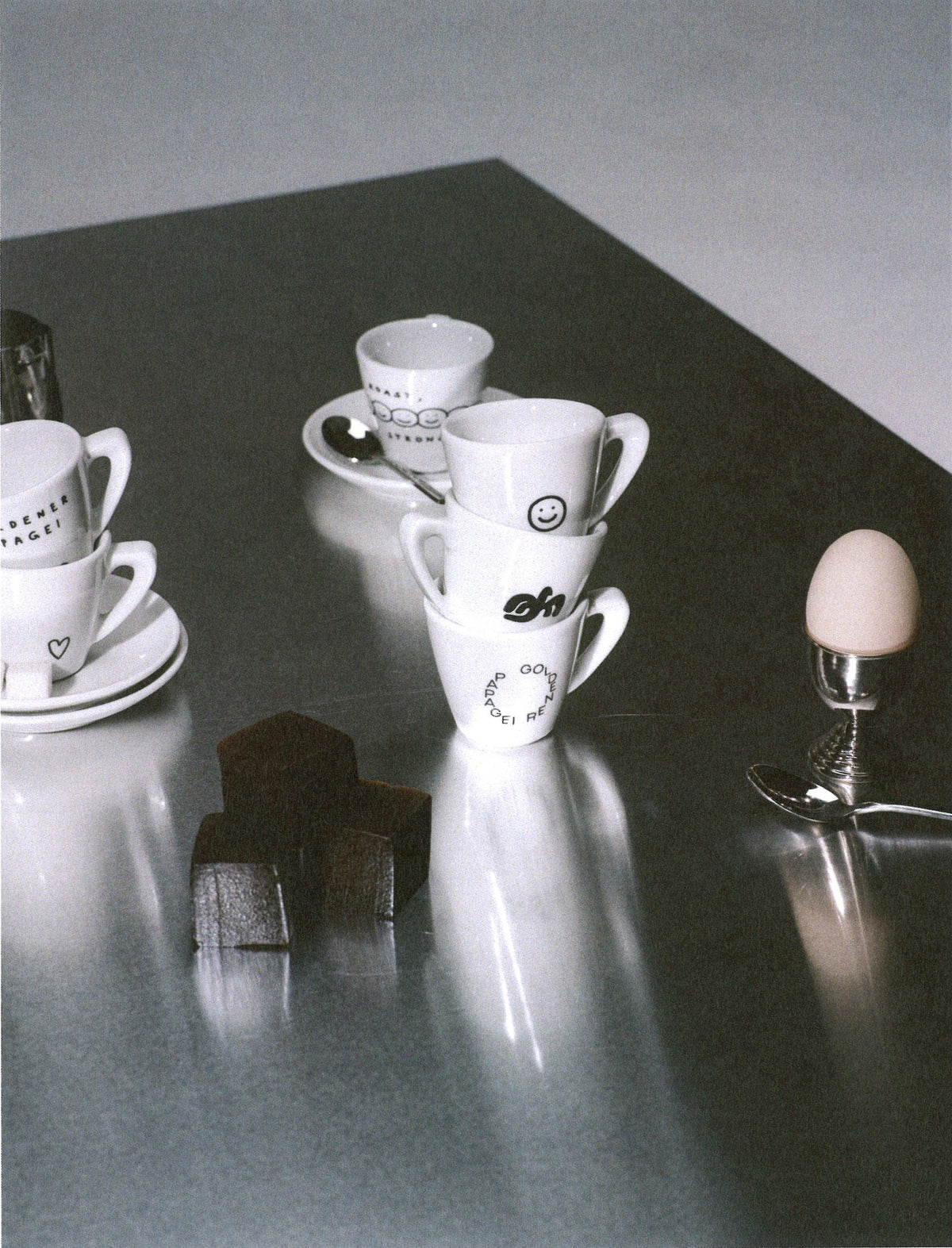 A square art print featuring a black and white photograph of white ceramic coffee cups and saucers on a reflective dark gray table, alongside a small egg on a stand and dark blocks, centered on a white background.