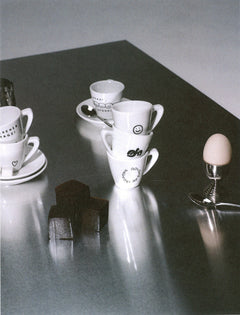 A square art print featuring a black and white photograph of white ceramic coffee cups and saucers on a reflective dark gray table, alongside a small egg on a stand and dark blocks, centered on a white background.